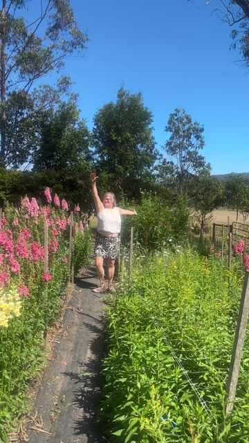 Person standing in lush garden with pink flowering plants and green vegetables, arms raised, clear blue sky.