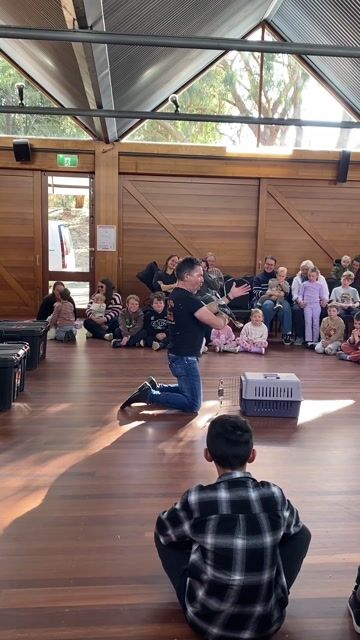 Instructor kneeling with a kookaburra while students sit and watch in a wooden hall with natural light