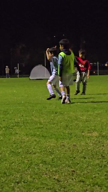 Young soccer players under field lights, one in green bib controlling the ball during evening training session.